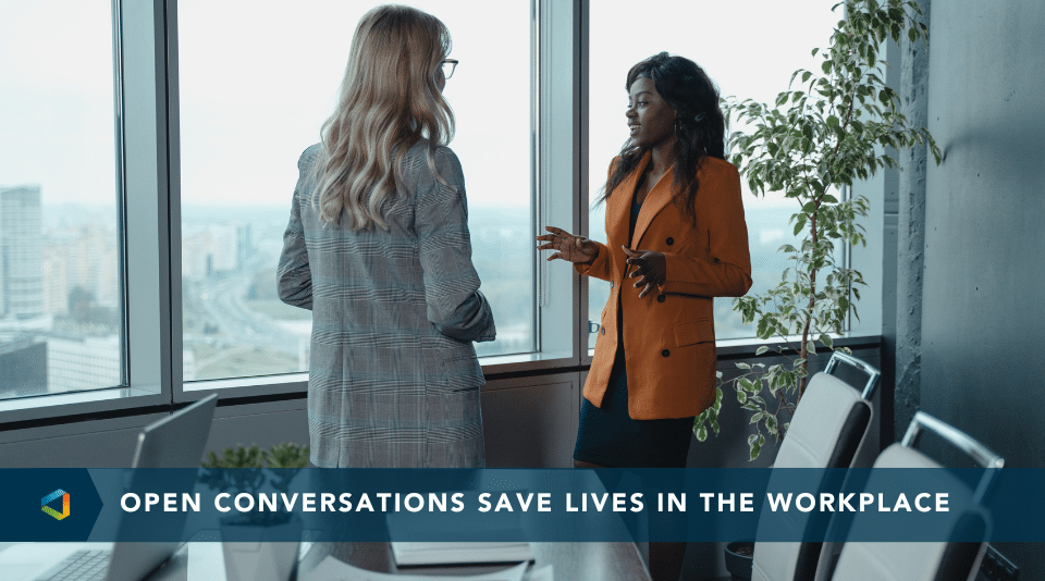 Two professional women in discussion by a large office window, symbolizing open workplace conversations about suicide prevention.